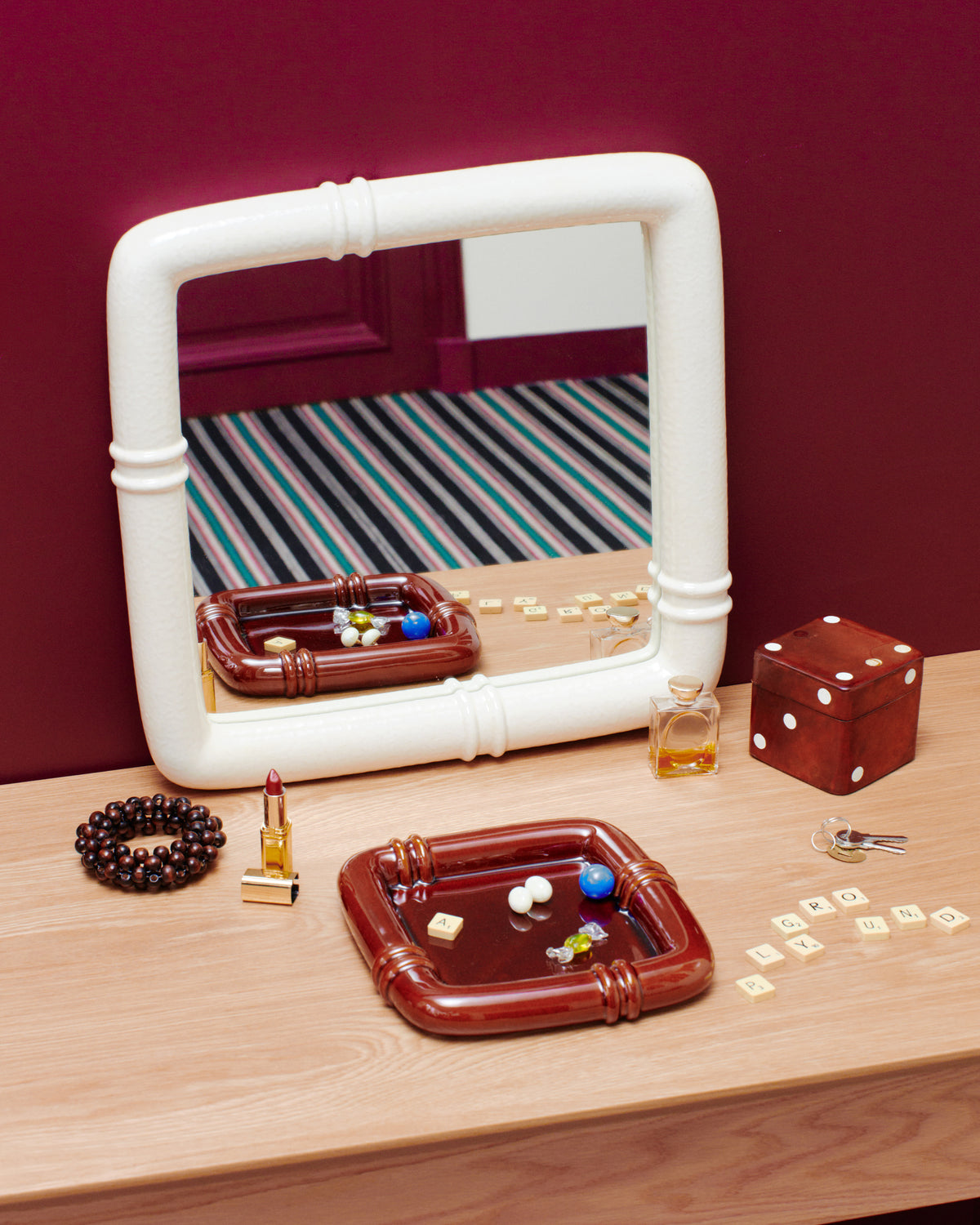Miroir Coco Ecru sur mur bordeaux, Vide-poches Coco Noisette garnis de billes, dé et bracelet