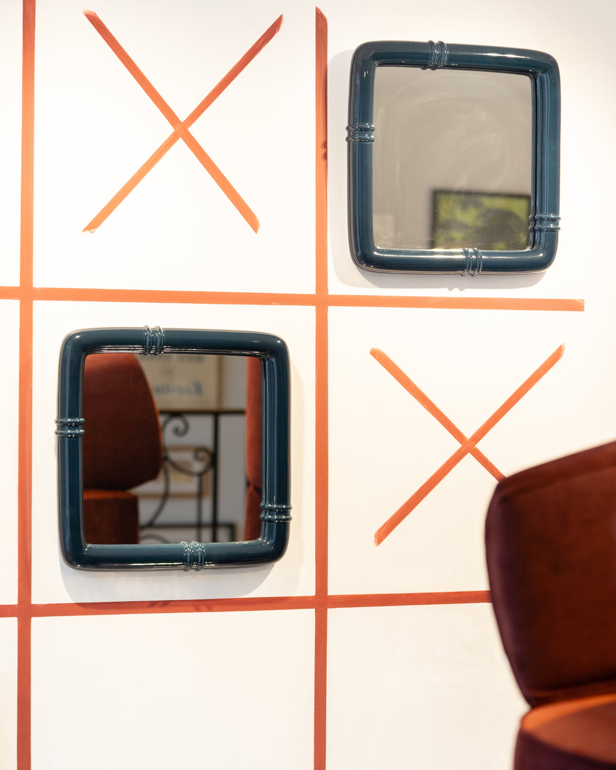 Miroirs Coco Bleu Noir accrochés en morpion sur un mur aux croix terracotta, pouf bordeaux